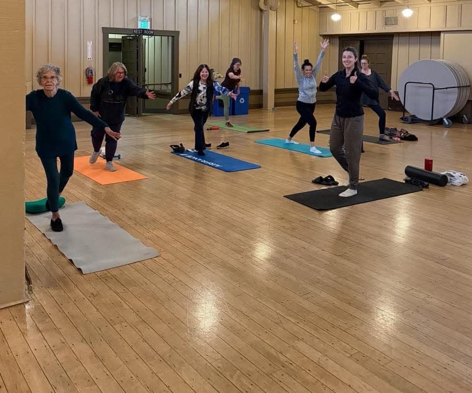 Group fitness class balancing on one leg on mats in a wood-floored community hall